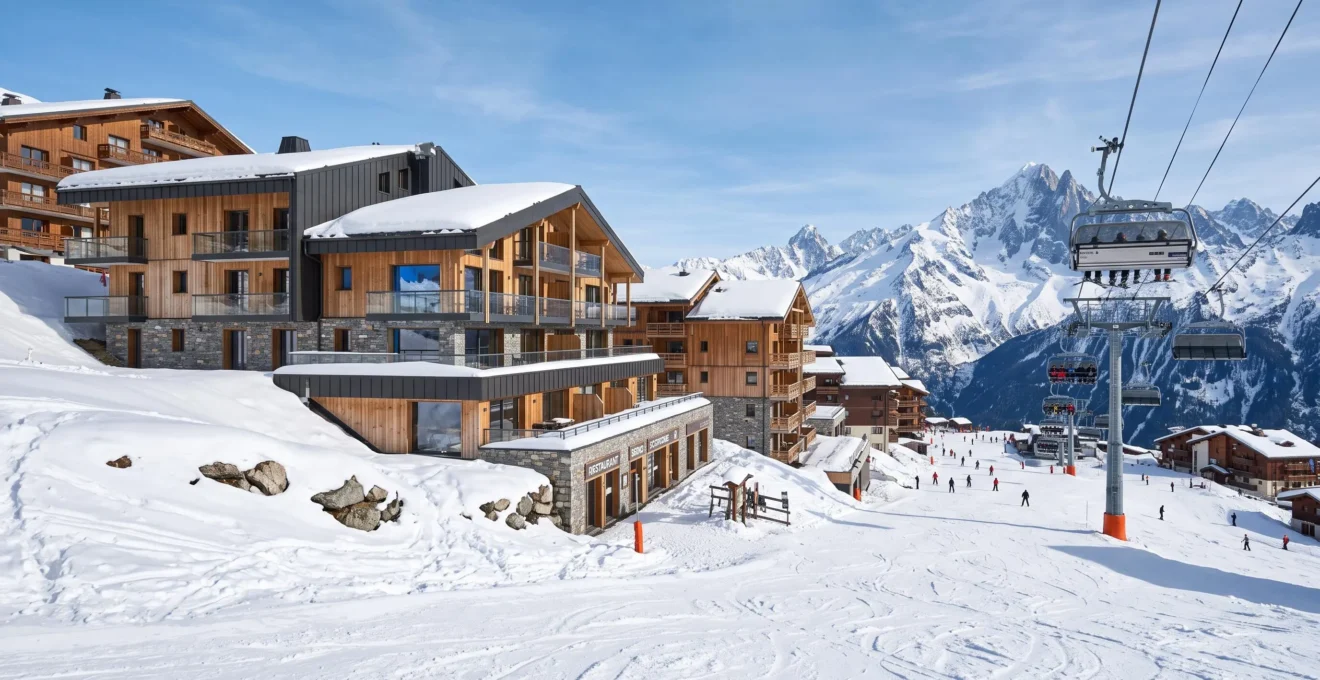 Vue en grand angle d'une station de ski alpine moderne en hiver avec des résidences contemporaines au pied des pistes, un télésiège visible en arrière-plan sous un ciel bleu lumineux
